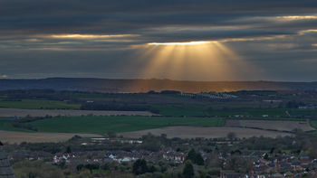 This landscape photograph captures a striking rural scene near Bolsover in Derbyshire, England, during the early evening in spring. The main focus is on the dramatic gods rays, also known as crepuscular rays, streaming through a break in the dense clouds and illuminating a solar farm set among the green fields. The rays of sunlight appear especially vivid as they descend from the sky, casting a golden glow over the array of solar panels and the surrounding countryside. The image highlights both the beauty of the natural lighting effects and the increasing use of renewable energy sources in the United Kingdom. In the foreground, clusters of homes are visible, while the rolling Derbyshire landscape extends into the distance beneath the dynamic cloud cover.